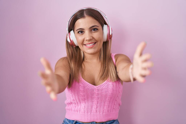 Jonge blonde vrouw luisteren naar muziek met behulp van koptelefoon glimlachend vrolijk aanbieden van handen geven van hulp en acceptatie.  - Foto, afbeelding