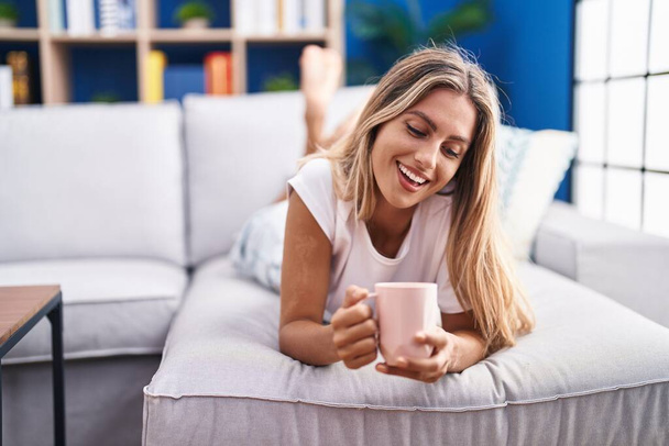 Young blonde woman drinking coffee lying on sofa at home - Photo, Image