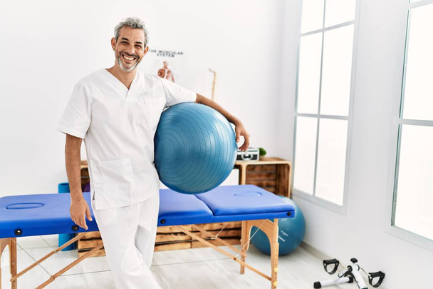 Középkorú ősz hajú férfi fizioterapeuta mosolygós magabiztos gazdaság jóga mat a rehab klinikán - Fotó, kép