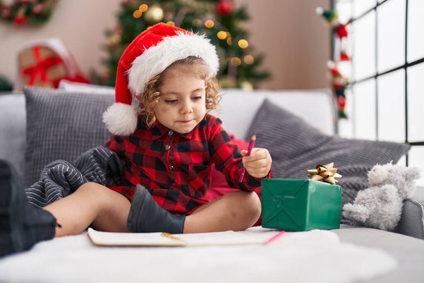 Adorable chica caucásica dibujando en el cuaderno sentado en el sofá por el árbol de Navidad en casa - Foto, Imagen