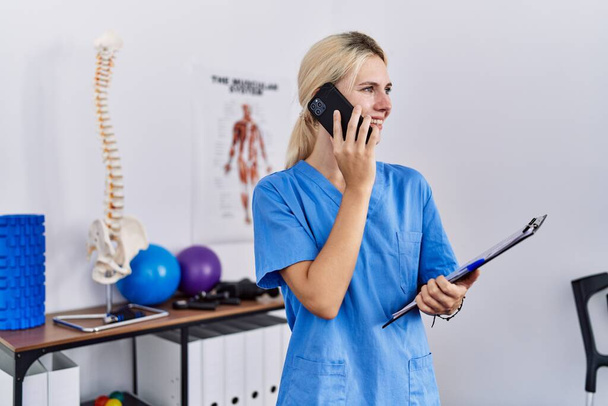 Young blonde woman pysiotherapist talking on smartphone holding clipboard at rehab clinic - Photo, Image