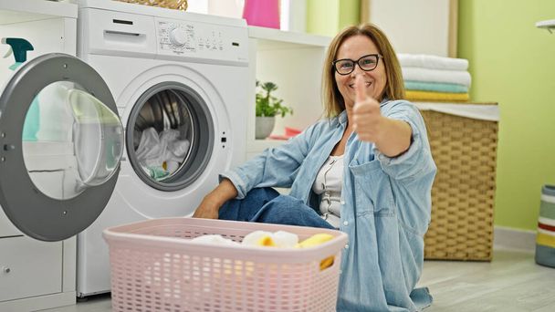 Medioevo donna ispanica sorridente fiducioso lavaggio vestiti facendo pollice verso l'alto gesto in lavanderia - Foto, immagini