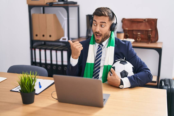 Knappe Spaanse man kijkt naar voetbalwedstrijd op kantoor duim wijzen naar de zijkant glimlachend blij met open mond  - Foto, afbeelding