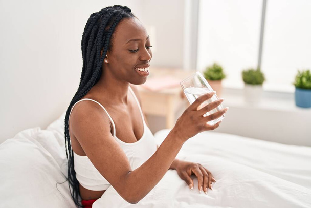 Αφροαμερικανή γυναίκα που πίνει νερό καθισμένη στο κρεβάτι στο υπνοδωμάτιο - Φωτογραφία, εικόνα