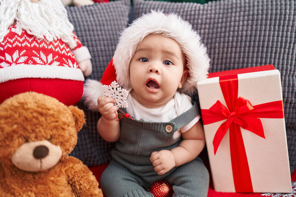 Entzückendes hispanisches Kleinkind, das zu Hause auf dem Sofa sitzt und Weihnachtsmütze trägt - Foto, Bild