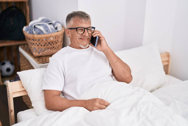 Hombre de pelo gris de mediana edad hablando en el teléfono inteligente sentado en la cama en el dormitorio - Foto, imagen
