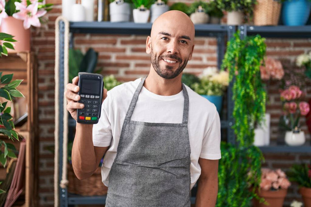 Ein Mann mittleren Alters mit Glatze, der im Blumenladen arbeitet, hält ein Datafon in der Hand und sieht im Stehen positiv und glücklich aus, lächelt mit einem selbstbewussten Lächeln und zeigt Zähne  - Foto, Bild