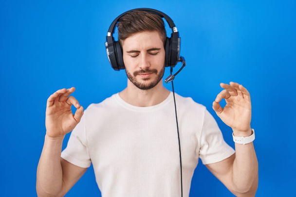 Ισπανόφωνος άνδρας με γενειάδα ακούγοντας μουσική φορώντας ακουστικά χαλαρός και χαμογελαστός με κλειστά μάτια κάνοντας χειρονομία διαλογισμού με δάχτυλα. έννοια γιόγκα.  - Φωτογραφία, εικόνα