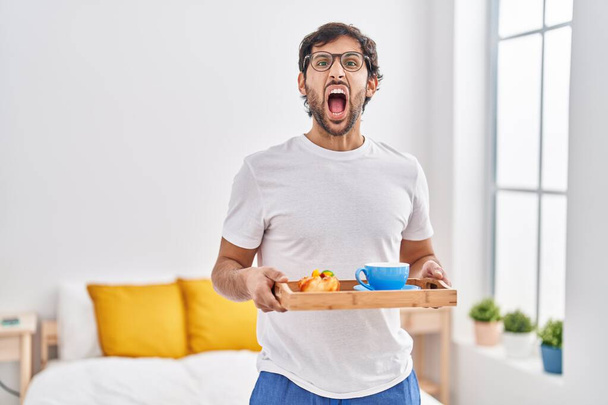 Pohledný latinskoamerický muž jedl snídani na posteli rozzlobený a šílený ječel, frustrovaný a zuřivý, křičel vzteky. vztek a agresivní koncept.  - Fotografie, Obrázek