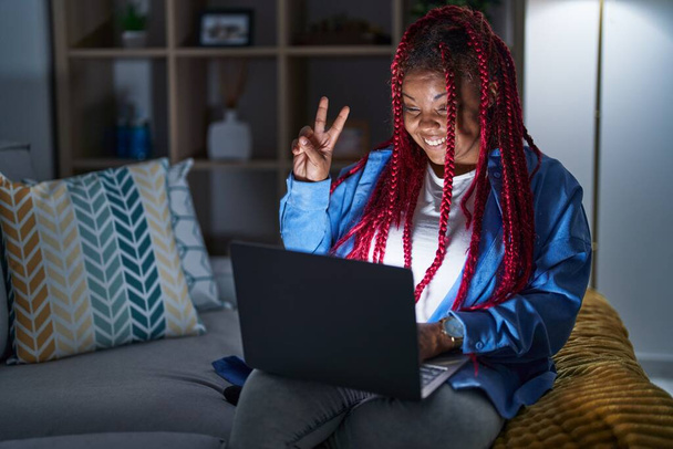 Mujer afroamericana con cabello trenzado usando computadora portátil por la noche sonriendo con la cara feliz guiñando el ojo a la cámara haciendo signo de victoria con los dedos. número dos.  - Foto, imagen