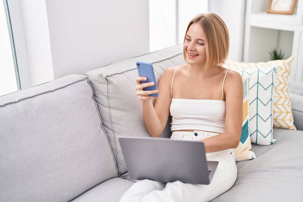Nuori blondi nainen käyttää kannettavaa tietokonetta ja älypuhelinta kotona - Valokuva, kuva