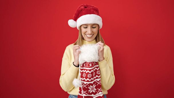 Молодая блондинка в рождественской шляпе смотрит внутрь носка на изолированном красном фоне - Фото, изображение