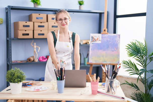Jonge blanke vrouw kunstenaar glimlachen zelfverzekerd met behulp van laptop in kunststudio - Foto, afbeelding