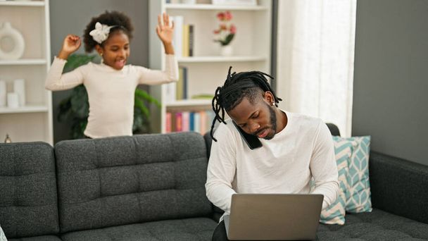 Le père et la fille afro-américains assis sur le canapé travaillent pendant que l'enfant dérange à la maison - Photo, image
