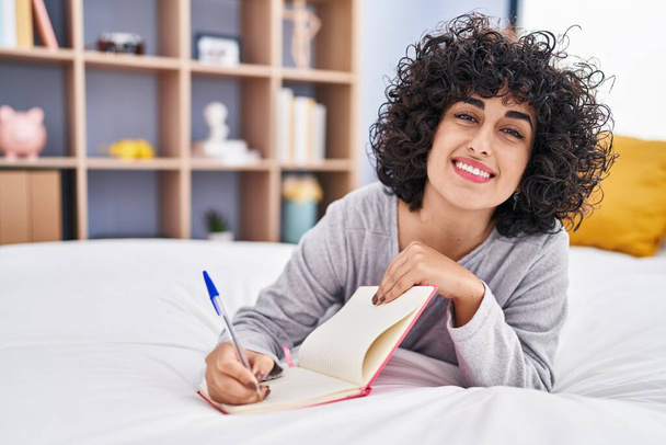 Junge Frau aus dem Nahen Osten schreibt auf Notizbuch, das im Schlafzimmer auf dem Bett liegt - Foto, Bild