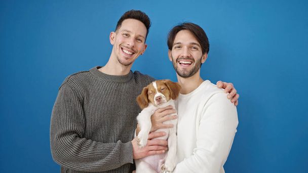 Двое мужчин улыбаются уверенно стоя обнимая собаку на изолированном синем фоне - Фото, изображение
