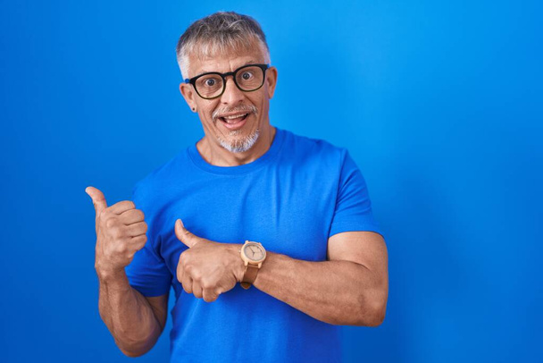 Hombre hispano con el pelo gris de pie sobre fondo azul apuntando hacia atrás con la mano y los pulgares hacia arriba, sonriendo confiado  - Foto, Imagen