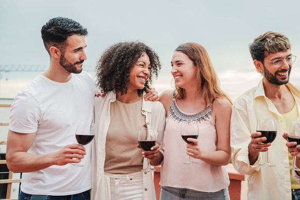 Group of young best friends having fun talking and drinking wine. Adult buddies smiling and holding wineglasses at party celebration. Men and women smiling with alcohol glasses at apartment rooftop - Photo, Image