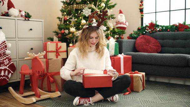 Fiatal szőke nő kicsomagolás ajándék ül a padlón karácsonyfa otthon - Fotó, kép
