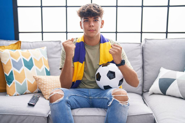 Ισπανόφωνος έφηβος κάθεται στον καναπέ βλέποντας ποδοσφαιρικό αγώνα ουρλιάζοντας υπερήφανος, γιορτάζει τη νίκη και την επιτυχία πολύ ενθουσιασμένος με σήκωσε τα χέρια  - Φωτογραφία, εικόνα