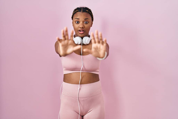 Africká americká žena s copánky na sobě sportovní oblečení a sluchátka dělá stop gesto s dlaněmi, naštvaný a frustrovaný výraz  - Fotografie, Obrázek