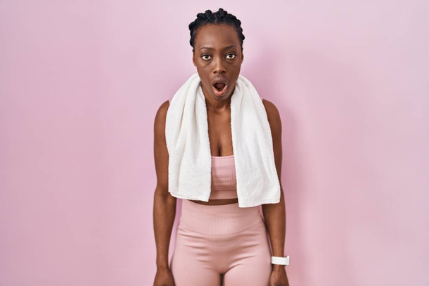 Piękna czarna kobieta ubrana w odzież sportową i ręcznik na różowym tle przestraszona i zszokowana zaskakującą ekspresją, strachem i podnieconą twarzą.  - Zdjęcie, obraz