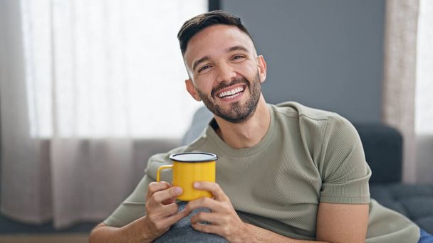 Young hispanic man drinking coffee sitting on sofa at home - Photo, Image