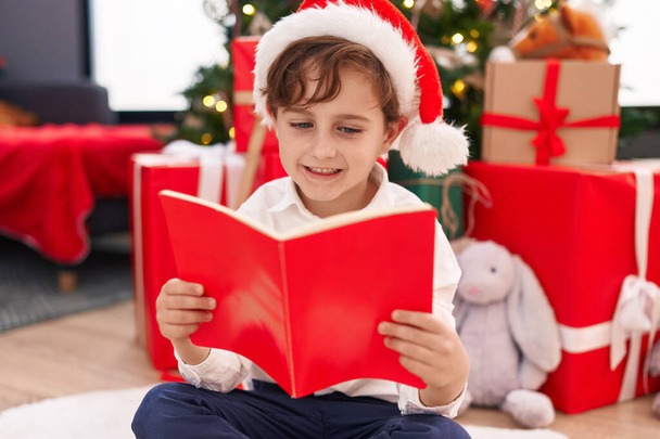 Suloinen latino poika lukeminen kirja istuu lattialla joulukuusi kotona - Valokuva, kuva