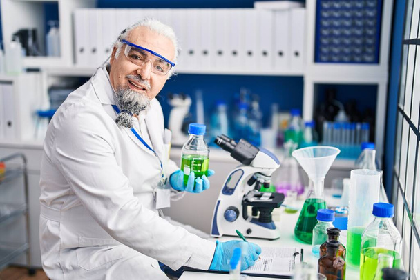 Středního věku šedovlasý muž vědec psaní na dokument drží láhev v laboratoři - Fotografie, Obrázek