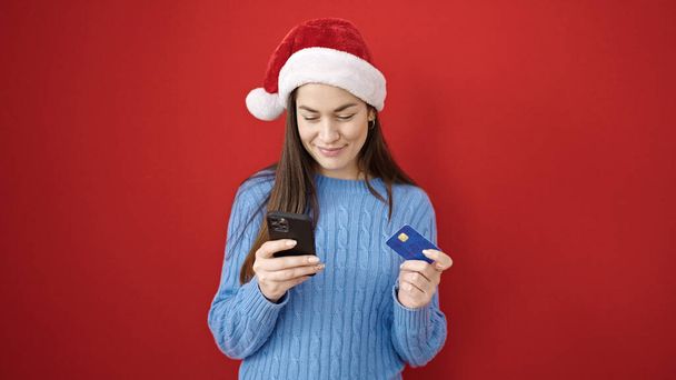 Νεαρή καυκάσια γυναίκα κάνει Χριστούγεννα online ψώνια με smartphone πάνω από απομονωμένο κόκκινο φόντο - Φωτογραφία, εικόνα