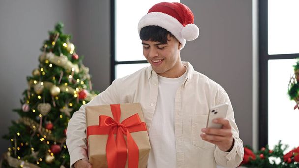Junger hispanischer Mann nutzt Smartphone mit Weihnachtsgeschenk zu Hause - Foto, Bild