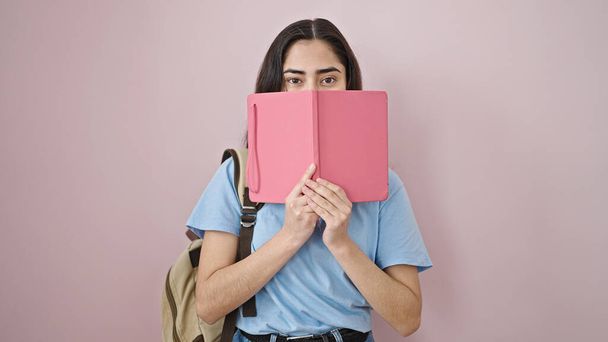 Junge schöne hispanische Studentin bedeckt Mund mit Buch über isoliertem rosa Hintergrund - Foto, Bild