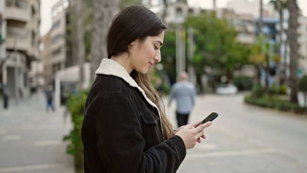 Молода красива іспаномовна жінка посміхається впевнено використовуючи смартфон на вулиці
 - Фото, зображення