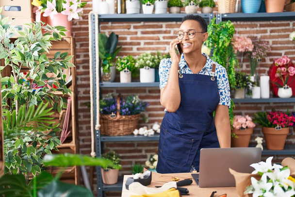 アフリカ系アメリカ人の女性の花屋は、花屋でラップトップを使用してスマートフォンで話して - 写真・画像