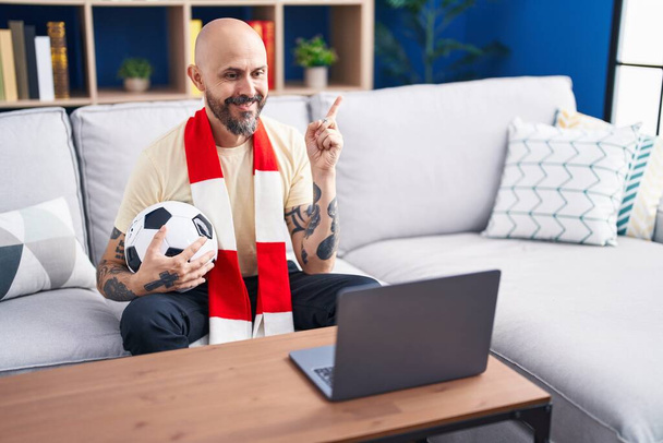 Hispánec s tetováním sledující fotbalový zápas chuligán držící míč na notebooku s úsměvem ukazující rukou a prstem na stranu  - Fotografie, Obrázek