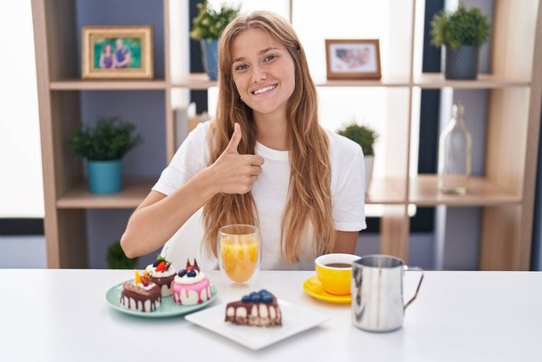 A fiatal kaukázusi nő süteményt eszik reggelire, boldog hüvelykujjával felemeli a kezét. jóváhagyása kifejezés nézi a kamera mutatja siker.  - Fotó, kép