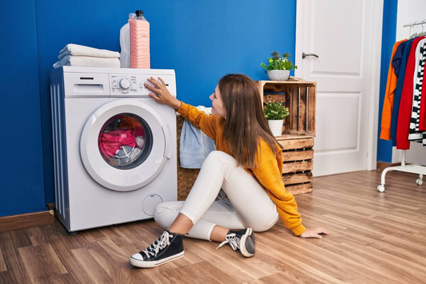 Mujer rubia joven sentada en el suelo girando la lavadora en la sala de lavandería - Foto, imagen
