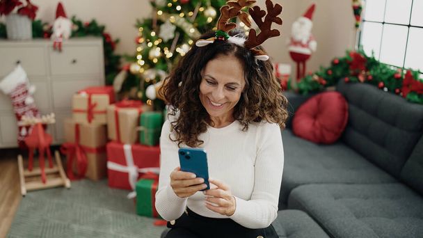 Keski-ikäinen latino nainen käyttää älypuhelinta istuu sohvalla joulupuulla kotona - Valokuva, kuva