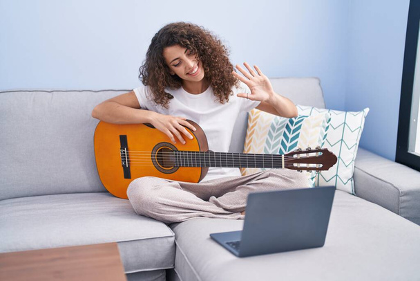 Young beautiful hispanic woman having online classical guitar class sitting on sofa at home - Fotó, kép