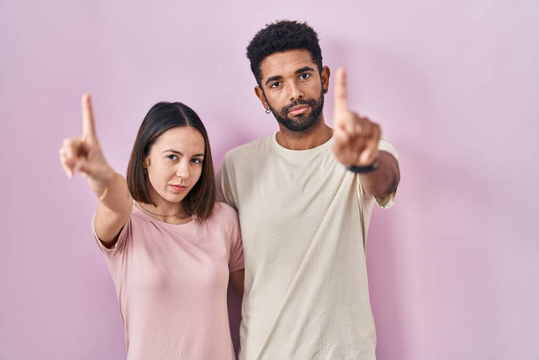 若いヒスパニックのカップルは,指で指を指し,怒った表情を指し,ジェスチャーを示さないピンクの背景の上に一緒に  - 写真・画像