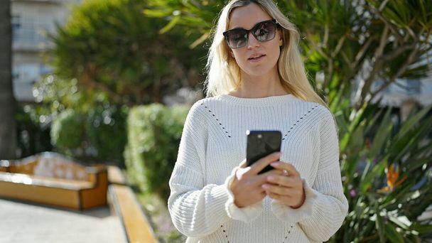Νεαρή ξανθιά γυναίκα που χρησιμοποιεί smartphone στέκεται με σοβαρή έκφραση στο πάρκο - Φωτογραφία, εικόνα