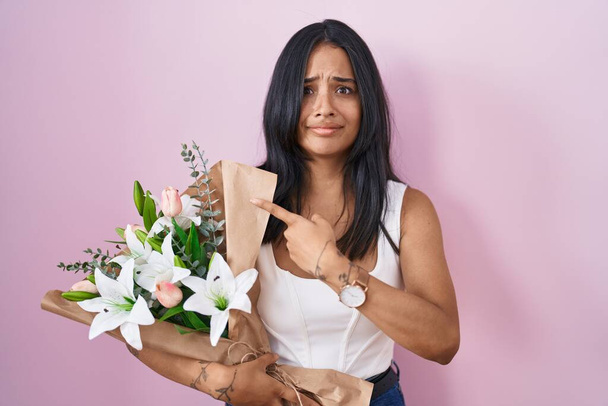 Mujer morena sosteniendo ramo de flores blancas señalando a un lado preocupado y nervioso con el dedo índice, preocupado y expresión sorprendida  - Foto, Imagen