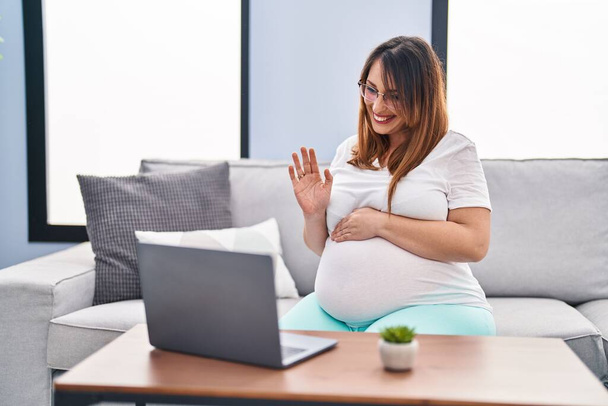 Jonge zwangere vrouw met video call zittend op de bank thuis - Foto, afbeelding