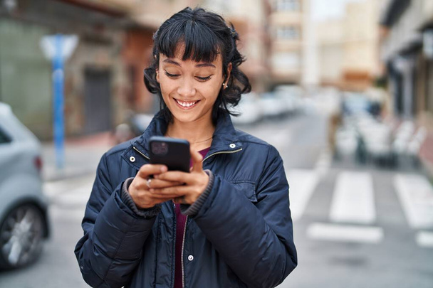 Genç, güzel, İspanyol bir kadın sokakta akıllı telefon kullanarak gülümsüyor. - Fotoğraf, Görsel