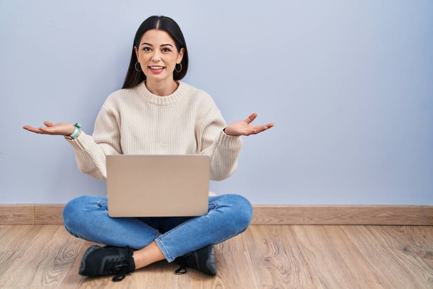 Giovane donna che utilizza il computer portatile seduto sul pavimento a casa sorridente allegro a braccia aperte come amichevole benvenuto, saluti positivi e fiduciosi  - Foto, immagini