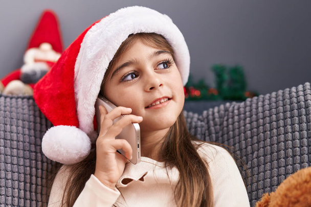Entzückendes hispanisches Mädchen mit Weihnachtsmütze spricht zu Hause auf dem Smartphone - Foto, Bild