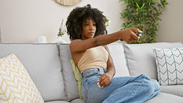 アフリカ系アメリカ人女性は,自宅で深刻な表現でソファーに座ってテレビを見ています - 写真・画像