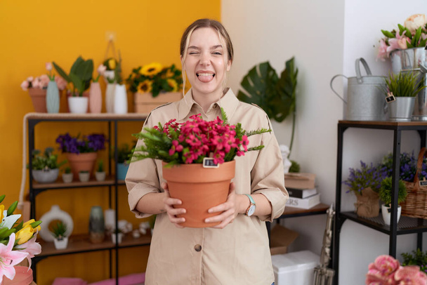 Jeune femme caucasienne travaillant à la boutique de fleuriste tenant pot plante langue collante heureux avec une expression drôle.  - Photo, image