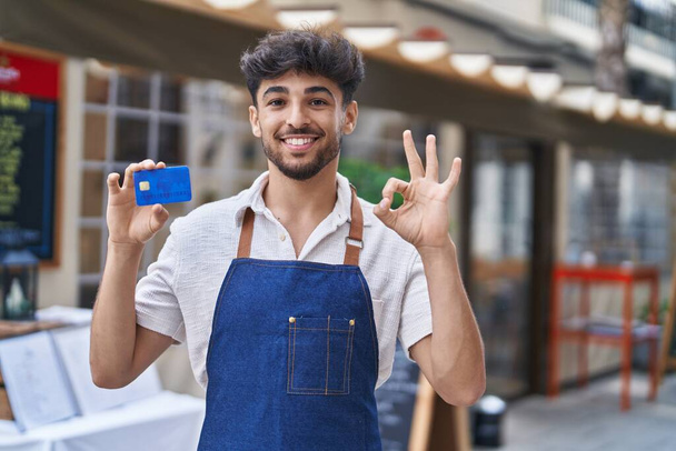 Hombre árabe con barba llevando delantal de camarero en la terraza del restaurante sosteniendo tarjeta de crédito haciendo signo bien con los dedos, sonriendo gesto amable excelente símbolo  - Foto, imagen