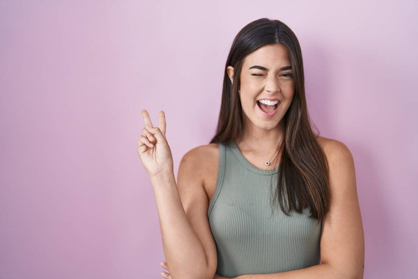 Hispanic woman standing over pink background smiling with happy face winking at the camera doing victory sign. number two.  - Photo, Image